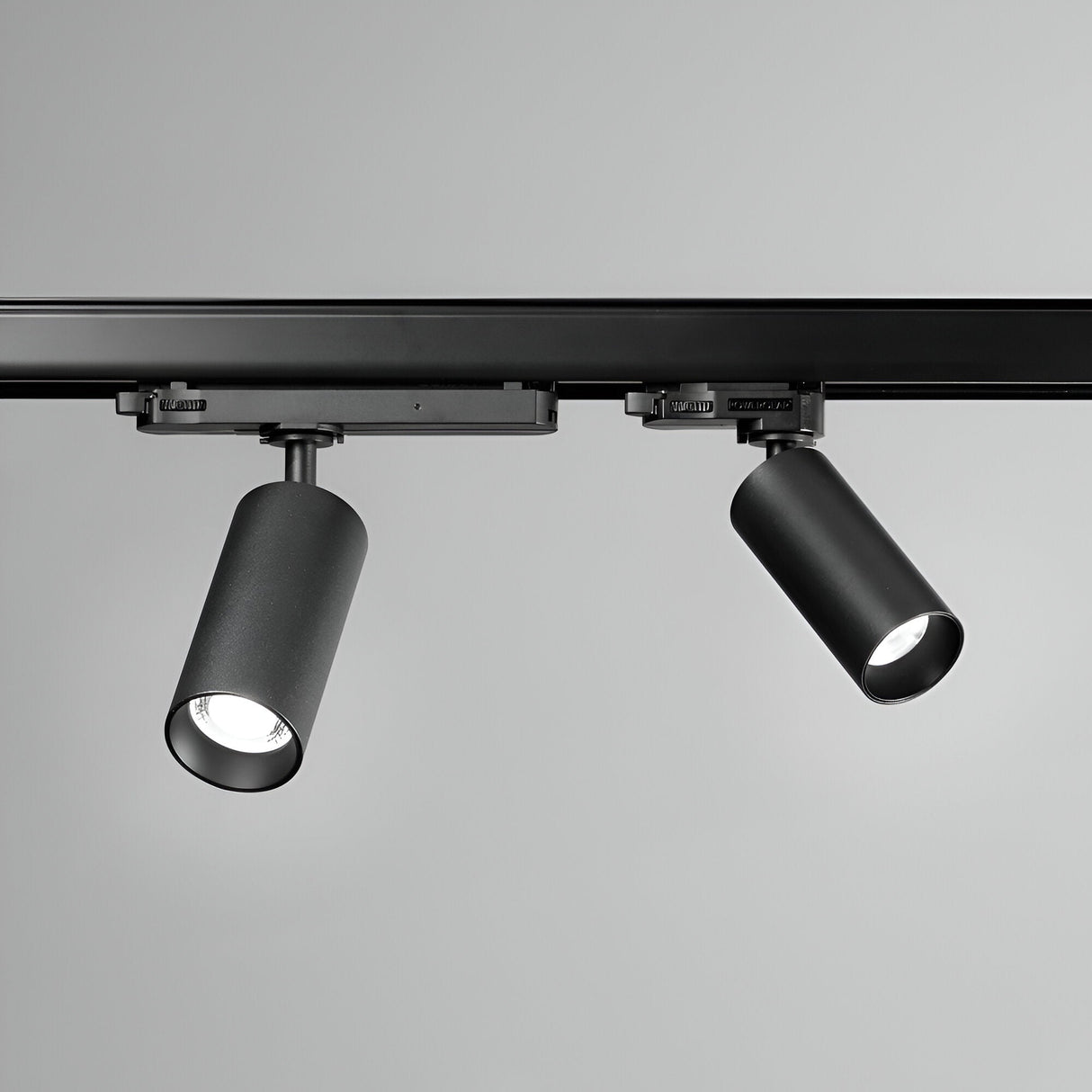 A pair of Glimmara LED Three Phase Track Spotlights, each with an 8W 3000K output in black, are mounted on a ceiling rail. One energy-efficient light is angled downward while the other features an adjustable head tilted slightly to the side, illuminating a plain grey background and highlighting their sleek, cylindrical design.