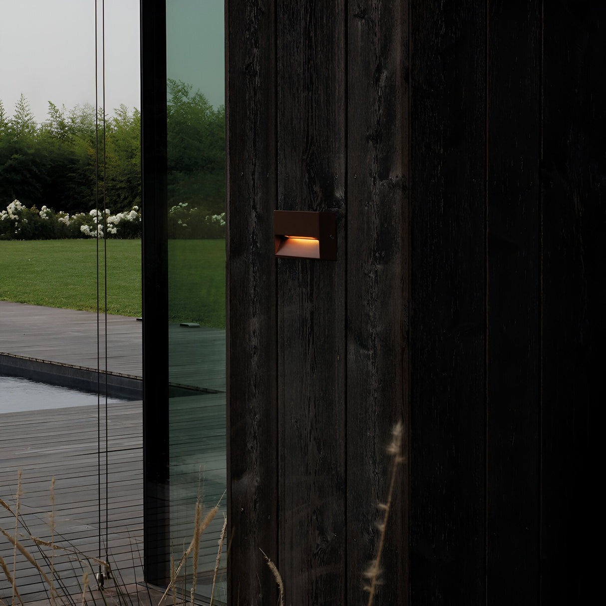 At dusk, the contemporary house exterior showcases a dark wooden wall bathed in the warm glow of the energy-efficient Arctis Small LED Outdoor Wall Light 1.5W 3000K - Brown, featuring an IP65 rating. The glass door mirrors the deck and a verdant garden filled with white flowers and shrubs.
