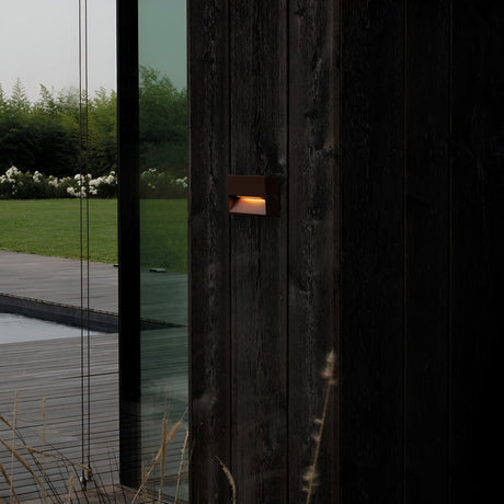 At dusk, the contemporary house exterior showcases a dark wooden wall bathed in the warm glow of the energy-efficient Arctis Small LED Outdoor Wall Light 1.5W 3000K - Brown, featuring an IP65 rating. The glass door mirrors the deck and a verdant garden filled with white flowers and shrubs.