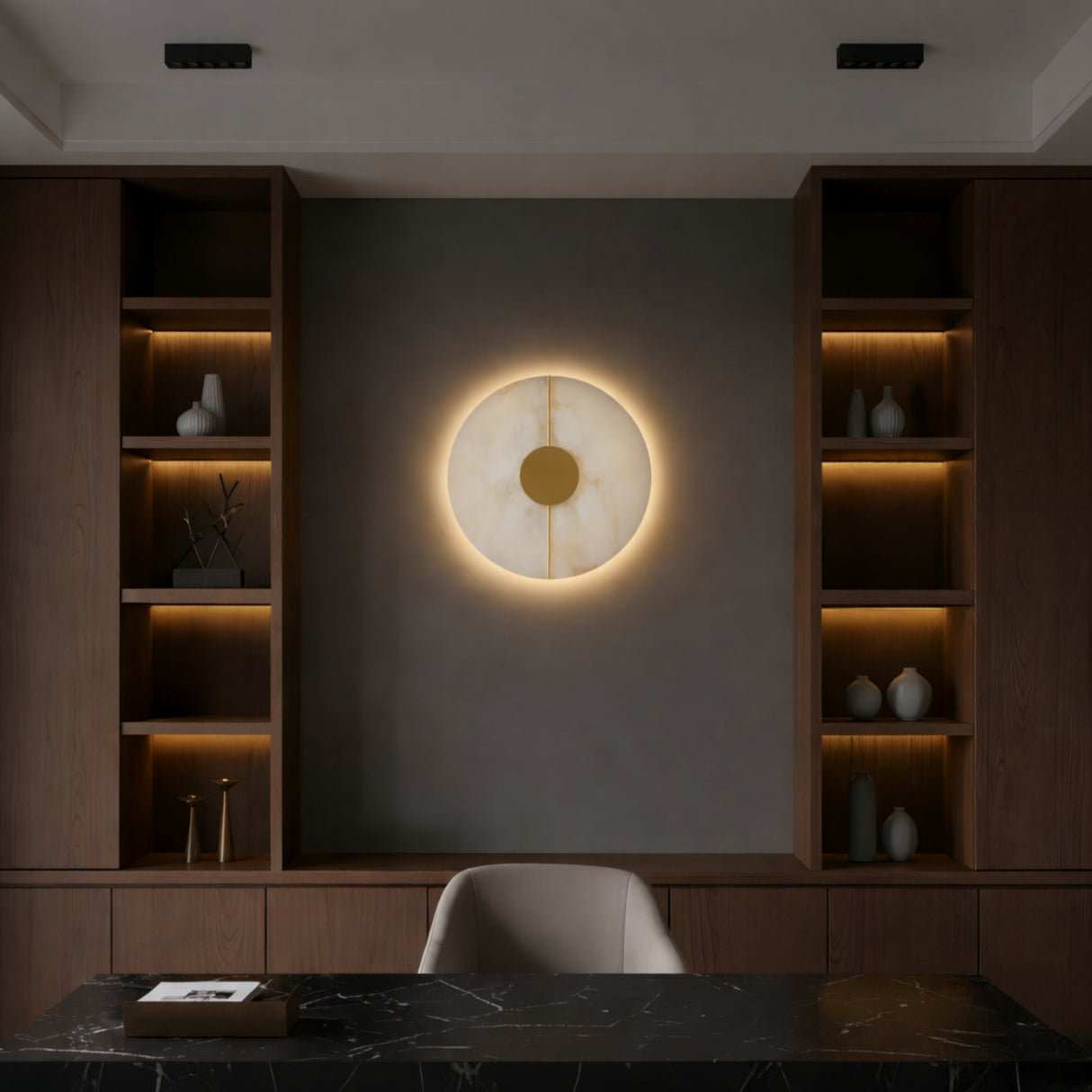 A modern office features dark wood shelves, a marble desk, a light gray chair, and the Elaris LED Round Alabaster Wall Light in soft white & brass centered on a gray accent wall, with vases and books decorating the shelves.