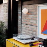 A modern interior features a turntable with a white record on top of a yellow cabinet. A framed abstract art piece and the Gleonis Wall Light in white, measuring 10cm, add charm to the brick wall. A leafy plant is partially visible on the left, enhancing the room's aesthetic.