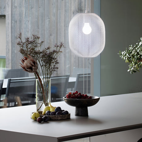 In a modern kitchen, the Aeron 1 Light Pendant in white, measuring 24cm, provides adjustable lighting over the countertop. On the counter, a black bowl holds grapes, apples, and plums next to a vase filled with assorted dried flowers. Through the window, there's a view of a patio featuring chairs with minimalist design.