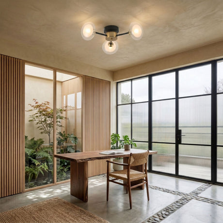 A modern office with a wooden desk and chair, large black-framed windows, indoor plants, stone floor accents, and the Galaxy 3 Light Wall/Semi-Flush Ceiling Light in Black & White with three round bulbs as the stylish centerpiece.