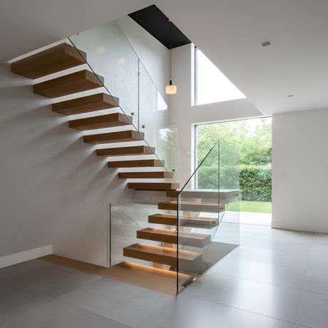 A modern floating wooden staircase with glass railings features the Draxen 1 Light Drum Alabaster Single Pendant Light in soft white and matte black, as large windows fill the minimalist space with natural light and views of greenery. Warm lighting highlights the stairs.
