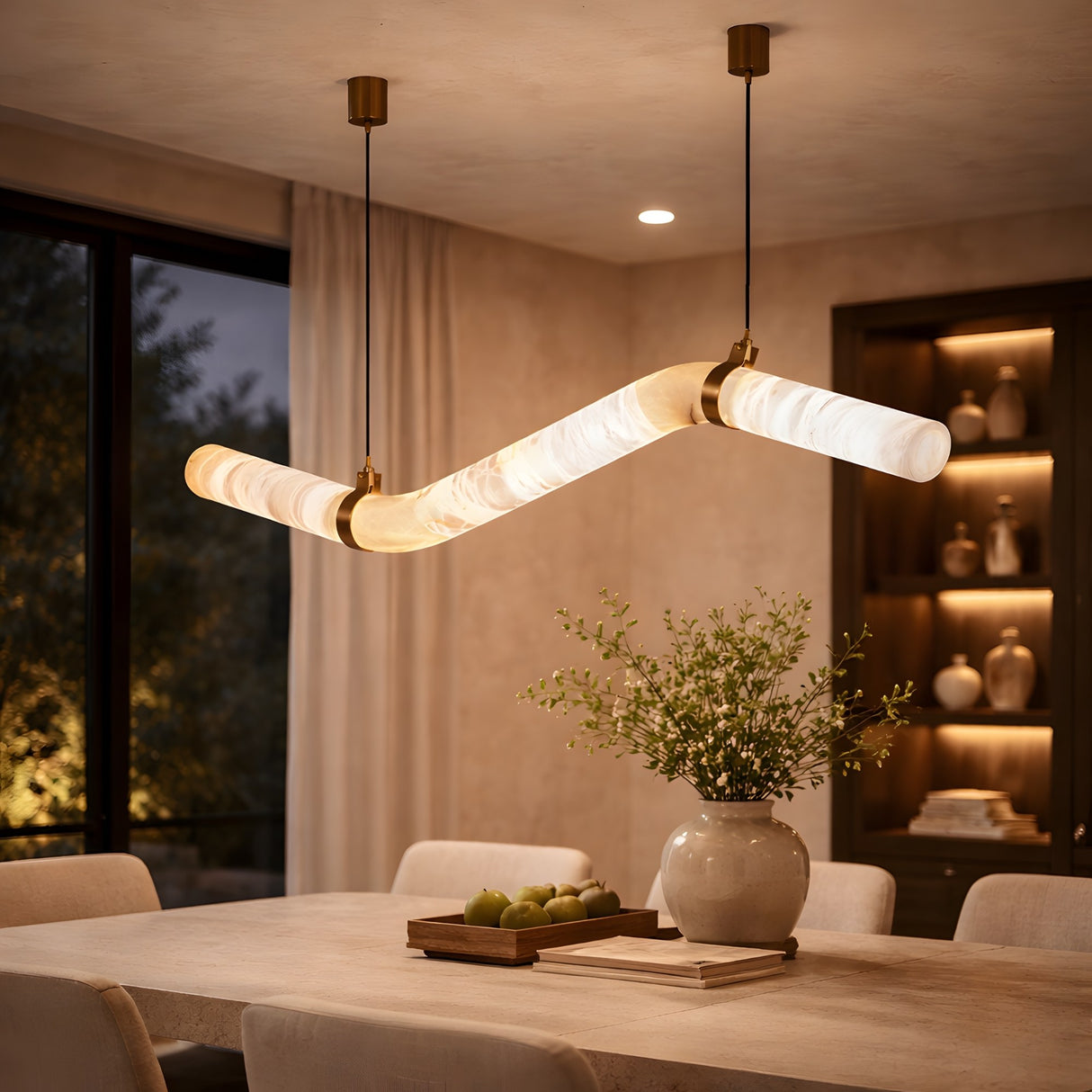 A modern dining room features the Zareon LED Small Articulated Alabaster Suspension Light – Soft White above a table decorated with a vase of greenery and apples, complemented by shelves with ceramics and large windows.