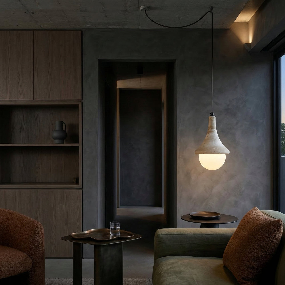 A modern living room with dark walls, cozy brown and green armchairs, the Velmora 1 Light Bell Travertine Single Pendant Light in light beige glowing overhead, built-in wooden shelves, a small table with a glass of water, and a hallway in the background.