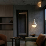 A modern living room with dark walls, cozy brown and green armchairs, the Velmora 1 Light Bell Travertine Single Pendant Light in light beige glowing overhead, built-in wooden shelves, a small table with a glass of water, and a hallway in the background.