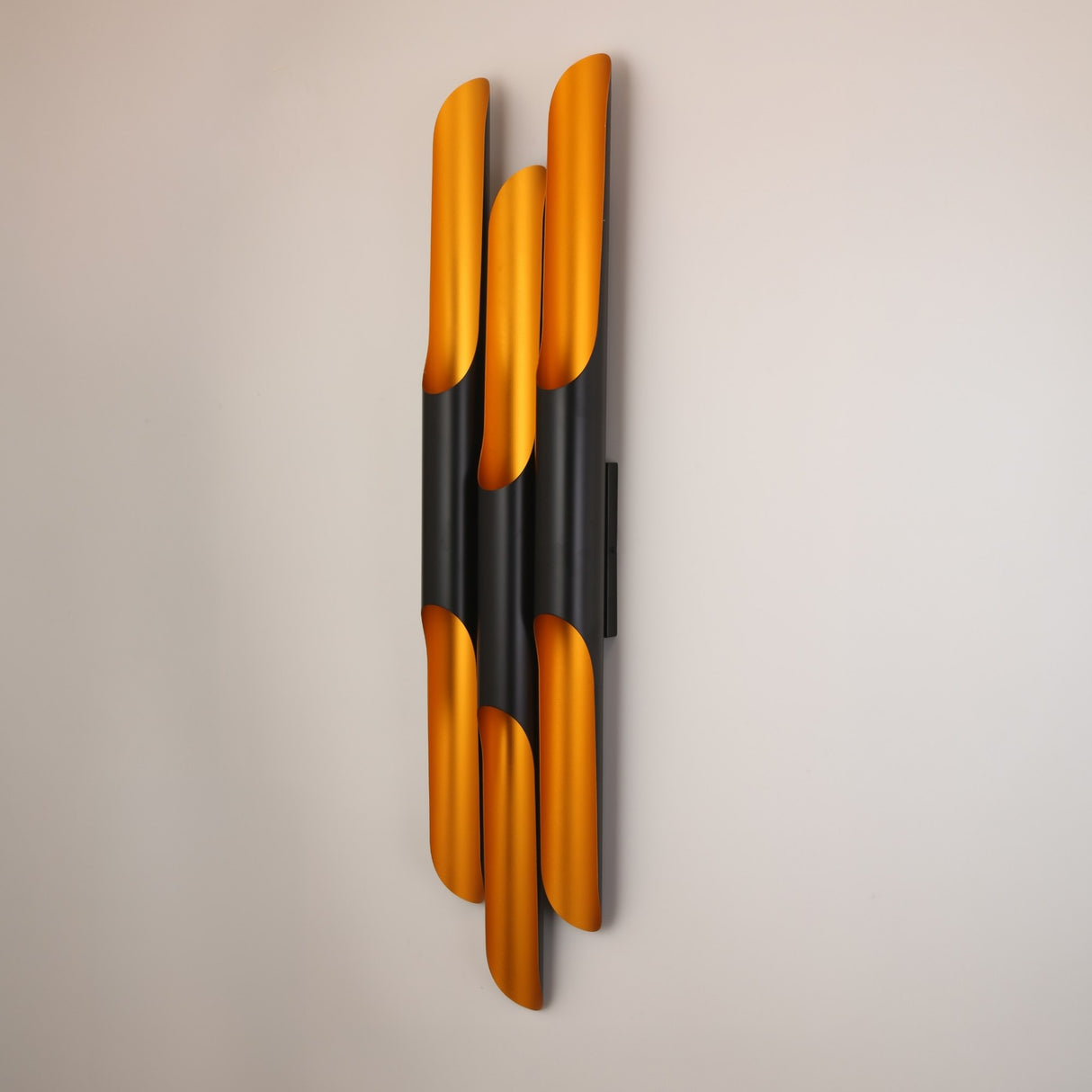 The Stealth 6 Light Wall Light in brass and black features vertical, overlapping metal pieces for striking, sculptural lighting when mounted on a light-colored wall.