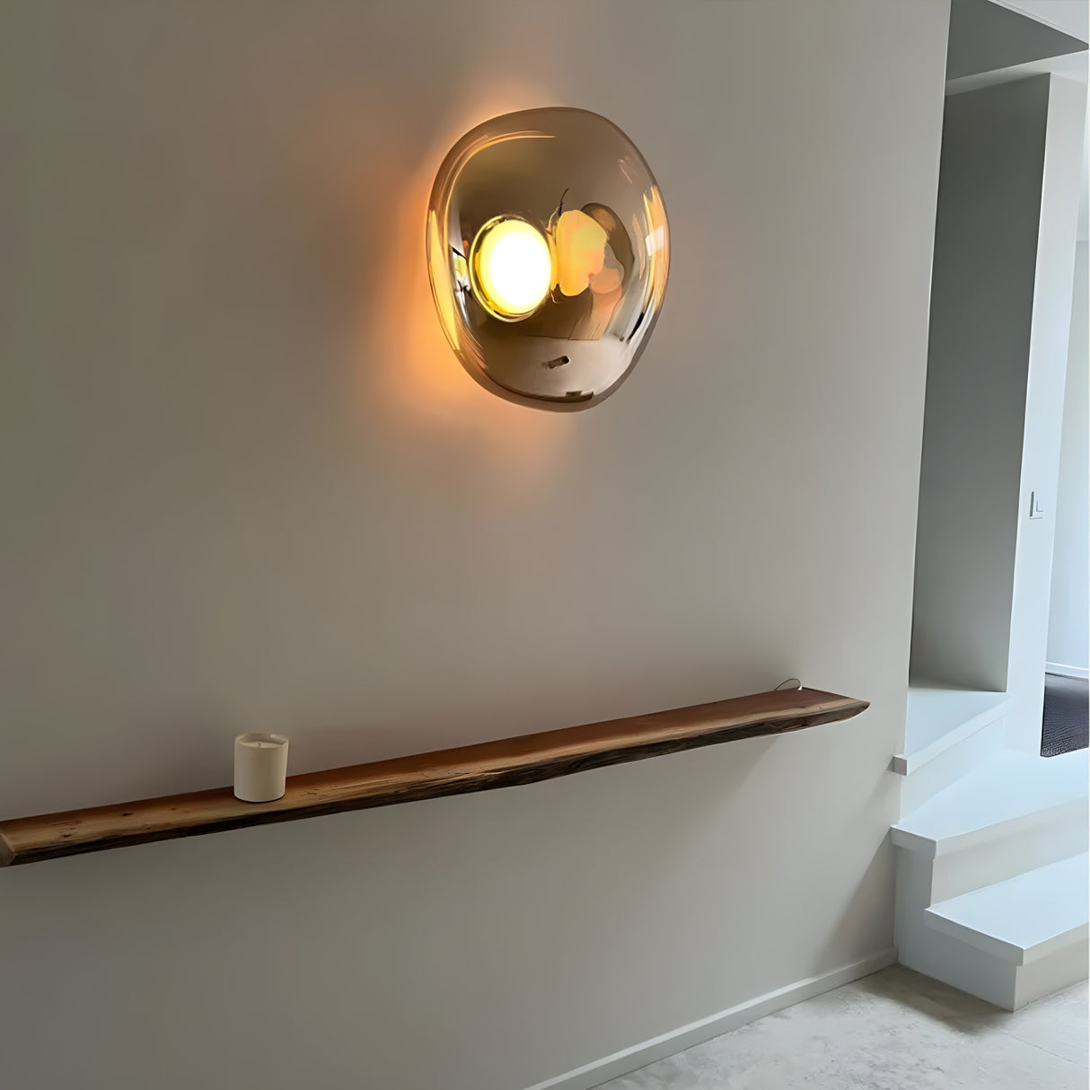 The Lava Large LED Wall & Ceiling Light in gold, with a rounded reflective glass cover, casts a warm glow on a minimalist wall above a wooden shelf with a white candle. White stairs appear to the right.