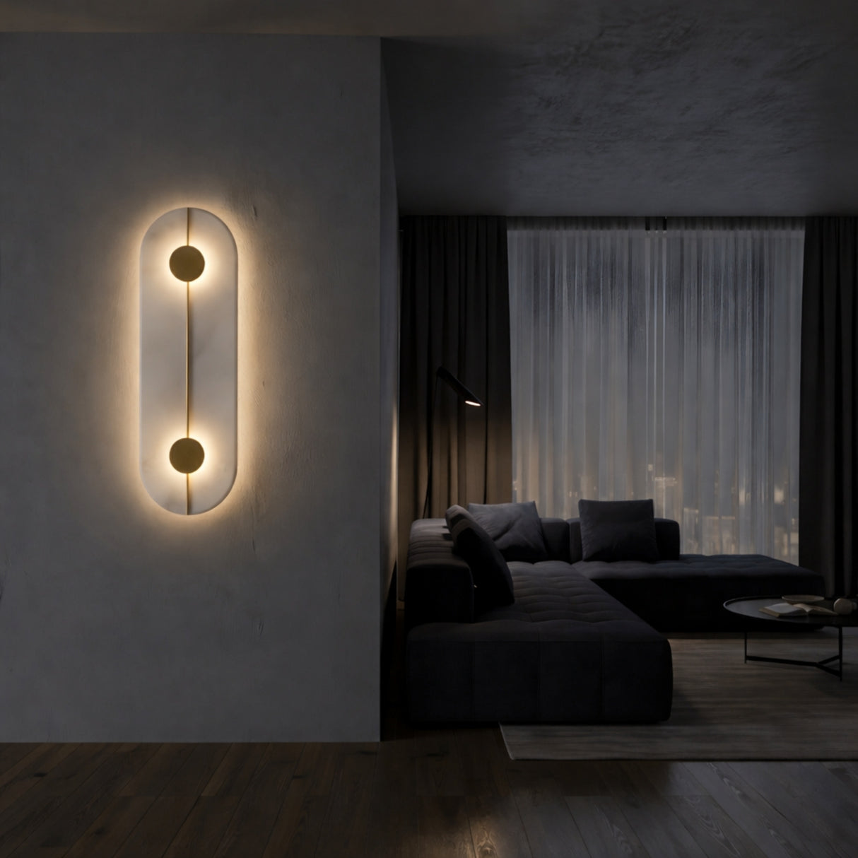 A modern, dimly-lit living room with dark furniture showcases the Elaris LED Capsule Alabaster Wall Light in soft white & brass, its sculptural twin circles glowing next to a large window with sheer curtains.