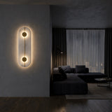 A modern, dimly-lit living room with dark furniture showcases the Elaris LED Capsule Alabaster Wall Light in soft white & brass, its sculptural twin circles glowing next to a large window with sheer curtains.