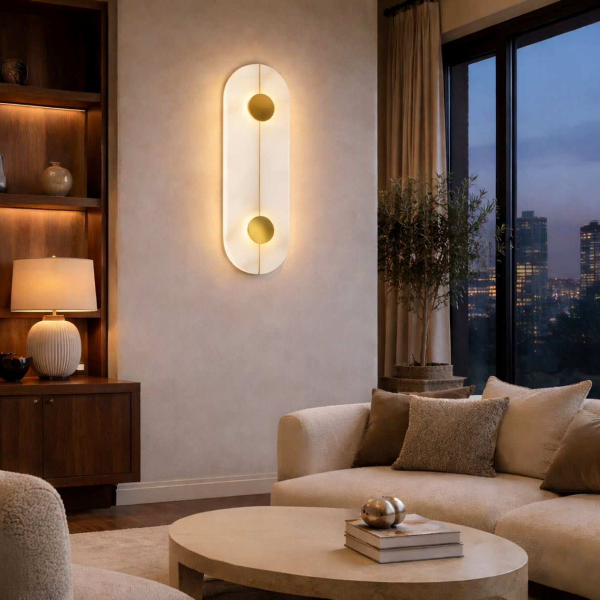 A modern living room with a round beige sofa, matching pillows, a circular coffee table, a wooden shelf with a table lamp, and the Elaris LED Capsule Alabaster Wall Light in Soft White & Brass. Large windows reveal a cityscape at dusk.