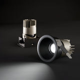 A close-up of a downlight with the Glistenis Trim Round Frame in black, emitting light against a dark background. Behind it, another similar downlight is unlit. This modern lighting installation showcases elegance and sophistication with the 77mm round frame.