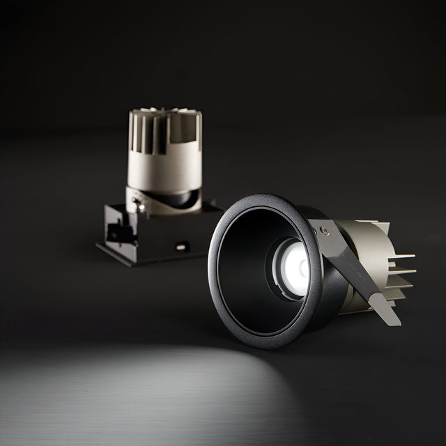 A close-up of a downlight with the Glistenis Trim Round Frame in black, emitting light against a dark background. Behind it, another similar downlight is unlit. This modern lighting installation showcases elegance and sophistication with the 77mm round frame.