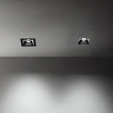 The image showcases a minimalist ceiling featuring two Glistenis Large Trimless Square Frames for Downlights in white. The downlights are switched on, casting a gentle illumination onto the gray surface below, enhancing the space with a modern and sleek atmosphere.