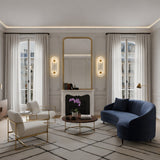 Elegant living room with tall windows, white curtains, a large wall mirror above the fireplace, Elaris LED Capsule Alabaster Wall Light in Soft White & Brass, navy curved sofa, two white chairs, round wooden coffee table, and geometric rug.