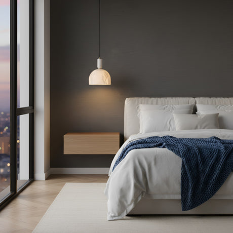 A modern bedroom featuring a large bed with white bedding and a blue throw, a wooden floating nightstand, and the Belvix 1 Light Small Dome Alabaster Single Pendant Light in Soft White & Brushed Brass by the window overlooking the city at sunset.