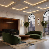 A modern lobby with marble walls, sculptural copper lighting, large arched windows, green velvet sofas set around a round wooden table, a patterned rug, potted plants, and Elaris LED Capsule Alabaster Wall Lights in soft white & brass.