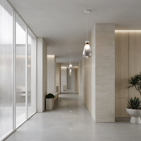 A modern hallway with light wood paneling, large frosted windows, potted plants, and neutral flooring features a bright, open feel enhanced by the Soffi Small Glass Pendant Light - Clear for a clean minimalist look.