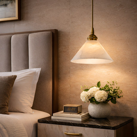 A cozy bedroom features a gray upholstered bed, white bedding, and a table with an alarm clock and white flowers. Above, the Novera 1 Light Conical Alabaster Pendant glows softly, framed by tan walls and partially visible artwork.