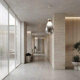 A modern, minimalist hallway featuring light wood paneling, large frosted glass windows, polished concrete floors, potted plants, and the Soffi Medium Glass Pendant Light - Clear hanging from the ceiling.