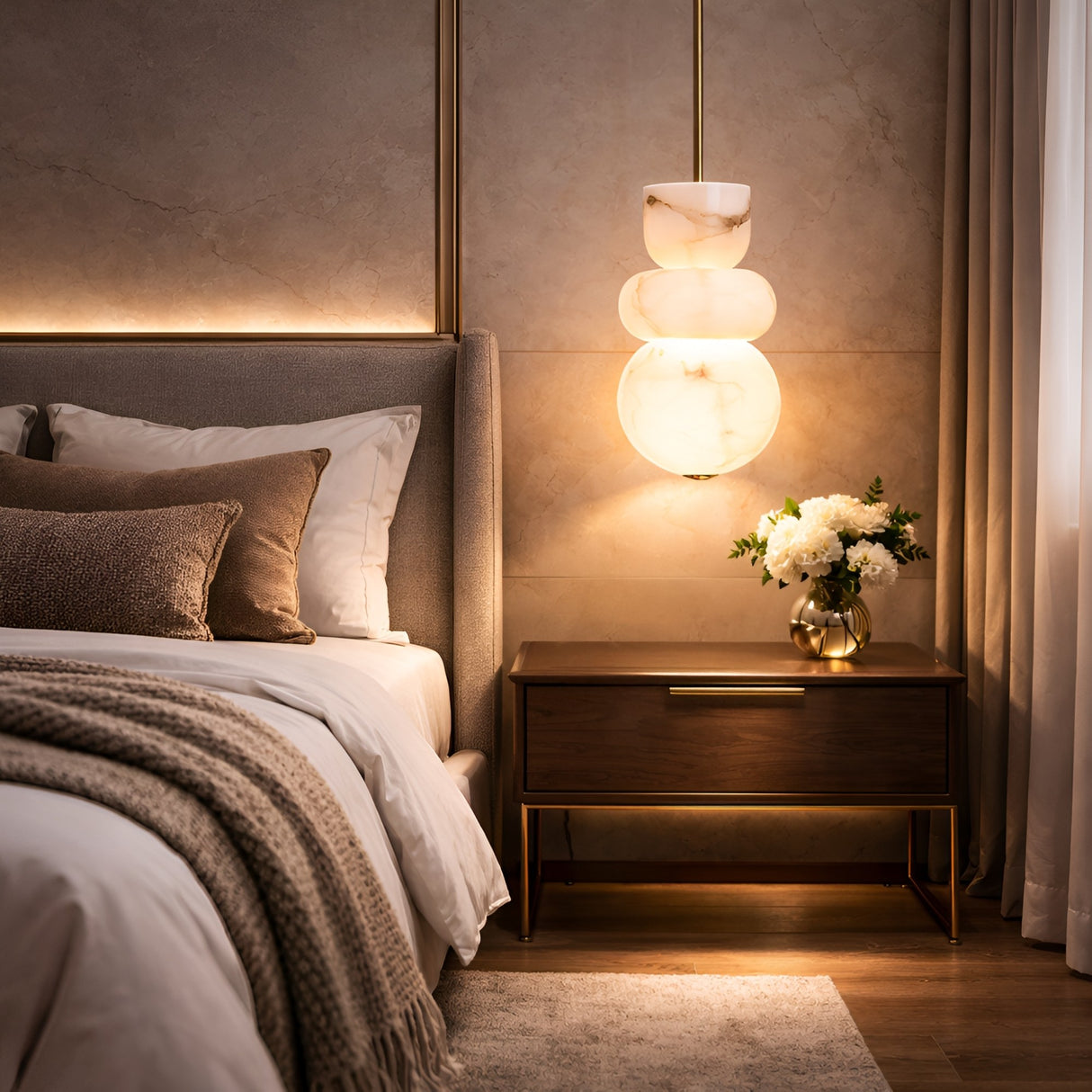 A cozy, modern bedroom with a neatly made bed, beige-brown bedding, a wooden nightstand with white flowers, and the Morvex LED Small Stacked Alabaster Single Pendant Light (Soft White & Brushed Brass) glowing beside soft curtains.