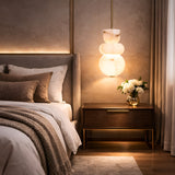 A cozy, modern bedroom with a neatly made bed, beige-brown bedding, a wooden nightstand with white flowers, and the Morvex LED Small Stacked Alabaster Single Pendant Light (Soft White & Brushed Brass) glowing beside soft curtains.