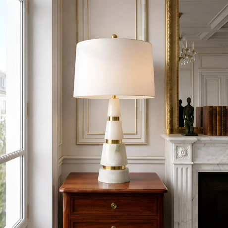The Zarethis 1 Light Stacked Alabaster Table Lamp in Brass & Soft White sits on a wooden chest by the window, adding elegance to a room with white paneled walls, a fireplace, books, and a small sculpture.