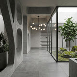 Minimalist interior with concrete walls and floors, large circular wall cutouts, and a striking Galaxy 10 Light Chandelier - Black & White above. A glass-enclosed courtyard holds a tree with mossy rocks; shelves are visible in the background.