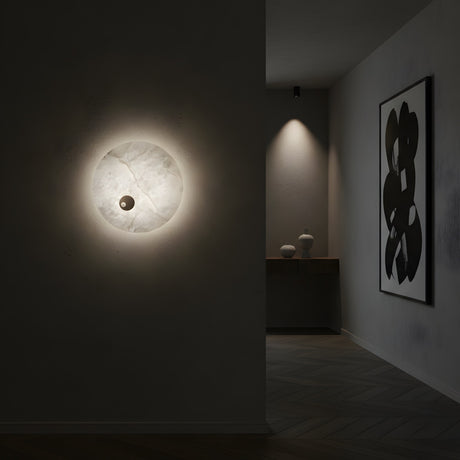 A minimalist interior features the Selvana LED Small Alabaster Wall Light - Soft White casting a gentle glow, an abstract black-and-white painting on the wall, and a console table with two vases in the background.