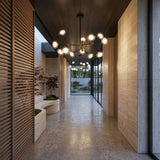 A modern hallway with stone walls, polished concrete floor, wooden slat panels, indoor plants in built-in planters, and the Galaxy 12 Light Chandelier - Black & White leading to large windows overlooking a landscaped garden.