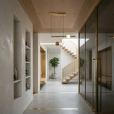 A modern, minimalist hallway with light wood and white walls features built-in shelves for vases, a Sottile LED Horizontal Suspension Light in gold above a glass partition with gold trim, and a floating staircase near a potted green plant. Natural light fills the space.