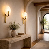 A softly lit hallway features arched doorways, a light stone console table with a ceramic vase of greenery, decorative bowls, and the Mica 1 Light Disc Alabaster Wall Light in Soft White & Brushed Brass, creating a warm, elegant atmosphere.