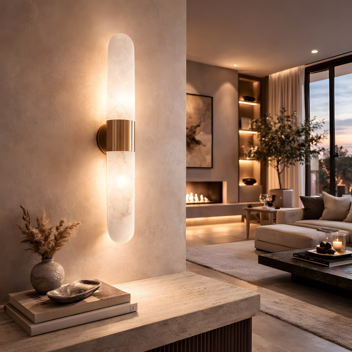 At sunset, a modern living room is illuminated by the Averin LED Alabaster Wall Light in Brushed Brass & Soft White, with neutral furniture, built-in shelves, a fireplace, decor accents, candles on the coffee table, and sheer curtained windows.