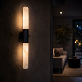 The Averin LED Alabaster Wall Light in Matte Black & Soft White features two cylindrical frosted lights, mounted vertically on a dark wall to softly illuminate a dim, minimalist living room.