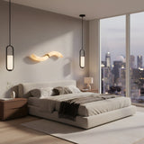 A modern bedroom with a large bed, neutral bedding, two wooden nightstands, the Eliona 1 Light Capsule Alabaster Single Pendant Light in matte black & soft white, a wavy wall lamp, and floor-to-ceiling windows overlooking the city at dusk.