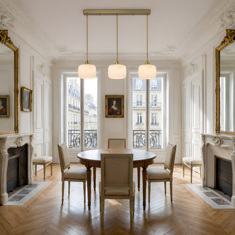 Elegant dining room with a round wooden table, six upholstered chairs, parquet wood floors, large windows, ornate mirrors, dual fireplaces, a wall portrait, and a Vantor 3 Light Linear Alabaster Bar Pendant Light in soft white & brushed brass above the table.
