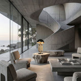 Modern concrete living room with large glass windows, minimalist furniture, a sculptural spiral staircase, and the Ascot 4 Light Small Table Lamp in Black & Gold on a geometric table. Natural light fills the spacious interior.