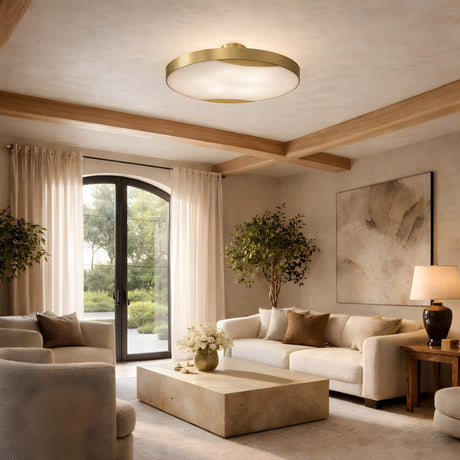 A modern living room with beige sofas, a wooden coffee table, large windows with sheer curtains, indoor plants, a table lamp, and the Marea Round LED Alabaster Semi-Flush Ceiling Light in brushed brass. Natural light enhances the cozy space.