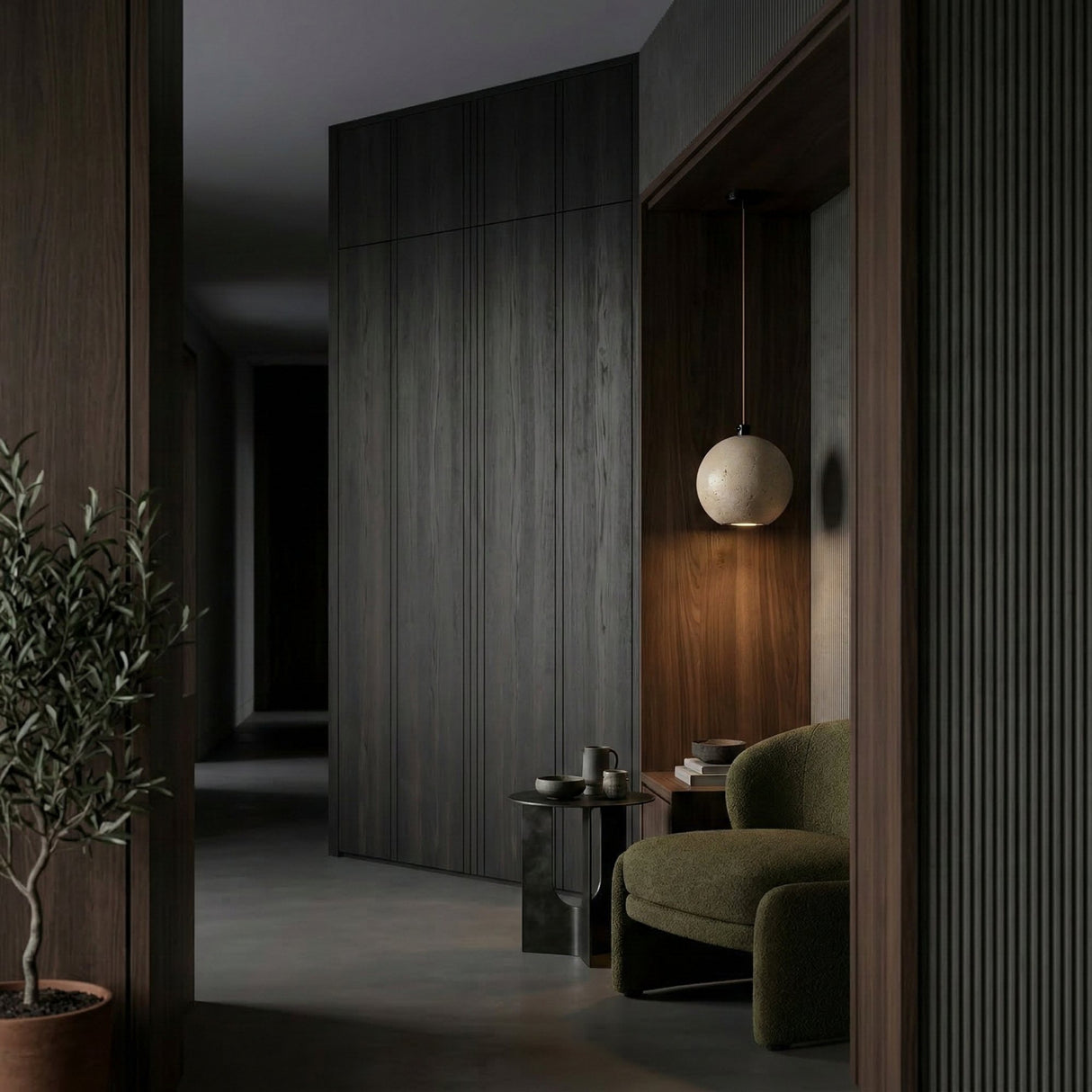 A modern, dimly lit interior features dark wood walls, a green armchair, books and cups on a side table, a potted plant, and the Orvello 1 Light Sphere Travertine Single Pendant Light (Light Beige), creating a cozy, minimalist atmosphere.
