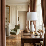 Elegant living room with a vintage armchair, marble fireplace, ornate mirror, and large windows with pink curtains. A wooden desk displays books, dried flowers, and the Calvero 1 Light Diamond Facet Marble Table Lamp in White & Brass. Sunlight fills the space.