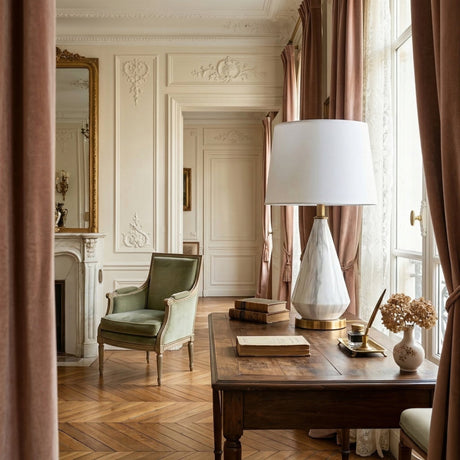 Elegant living room with a vintage armchair, marble fireplace, ornate mirror, and large windows with pink curtains. A wooden desk displays books, dried flowers, and the Calvero 1 Light Diamond Facet Marble Table Lamp in White & Brass. Sunlight fills the space.