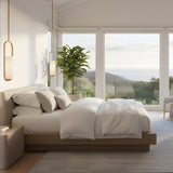 Eliona Capsule alabaster pendant lights in brushed brass hanging in a bright bedroom with white bedding, wooden bed frame, and coastal view windows.
