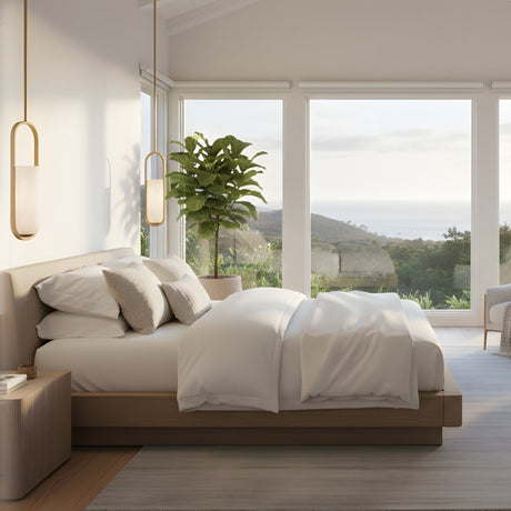 Eliona Capsule alabaster pendant lights in brushed brass hanging in a bright bedroom with white bedding, wooden bed frame, and coastal view windows.