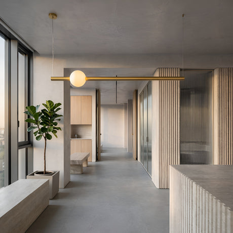 Minimalist interior with neutral tones, large windows, a potted plant, light wood accents, and the Sottile LED Horizontal Suspension Light in gold. Textured walls and built-in seating complete this bright, open contemporary space.