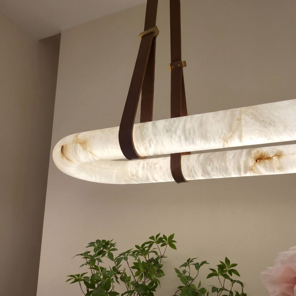 Cintura LED Small Alabaster Floating Pendant Light in soft white, showing the illuminated oval stone ring held by brown leather straps and copper fittings, in a room setting with plants below.