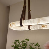 Cintura LED Small Alabaster Floating Pendant Light in soft white, showing the illuminated oval stone ring held by brown leather straps and copper fittings, in a room setting with plants below.