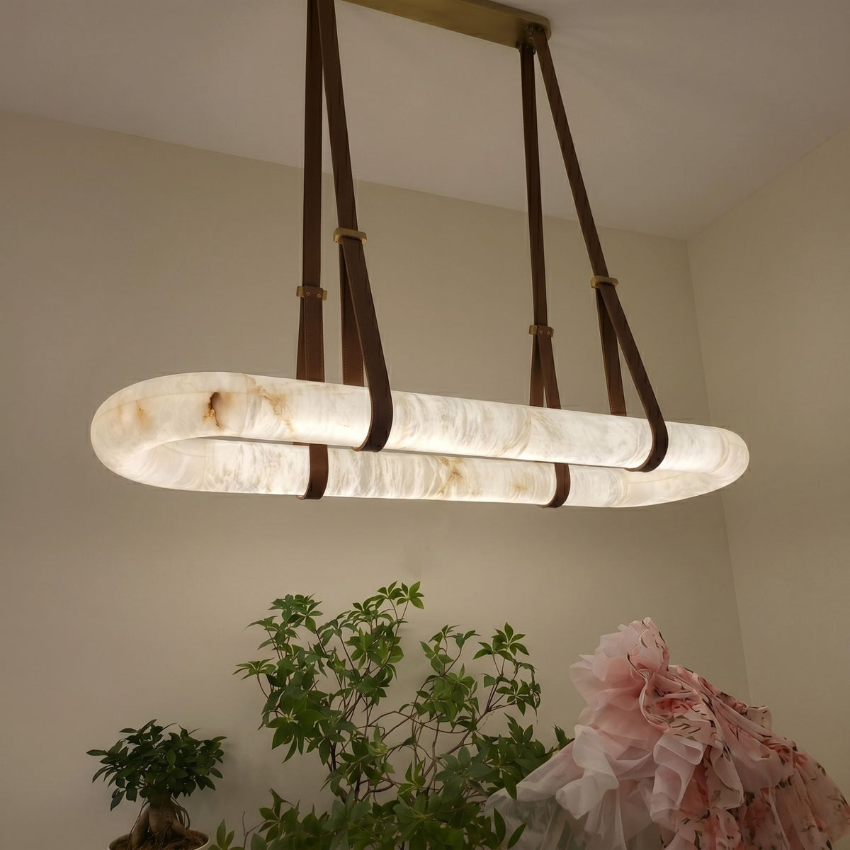 Cintura LED Small Alabaster Floating Pendant Light in soft white, an oval alabaster ring suspended by brown leather straps from a copper ceiling bar, shown illuminated above greenery in a dining room.