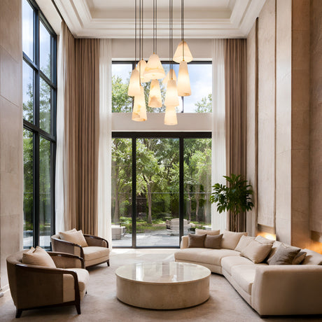 A modern living room with high ceilings, tall windows, neutral-toned furniture, a round marble coffee table, the Sorrelis 9 Light Cluster Alabaster Pendant in Soft White & Brushed Brass, and views of a lush green garden.