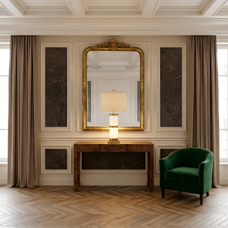 Elegant room with a large gold-framed mirror above a wooden console table featuring the Aurelin 1 Light Column Alabaster Table Lamp in Brass & Soft White, dark marble wall insets, beige curtains, and a green velvet armchair on herringbone wood floors.